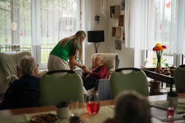 A caregiver offers a drink to an elderly woman sitting in a cozy armchair indoors.