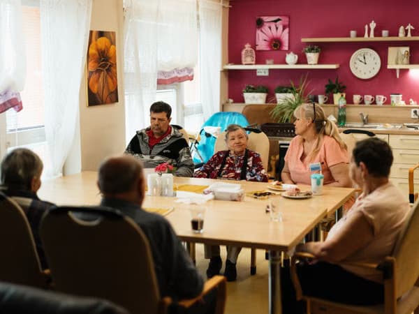 Group of older adults and caregivers conversing around a table in a bright room.