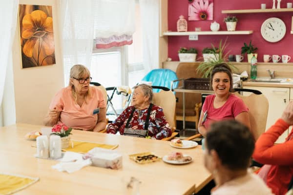Three women of different ages, including a caregiver, share snacks and conversation around a table.