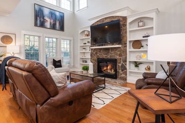 A cozy living room features brown leather sofas, a stone fireplace, and built-in white shelves.